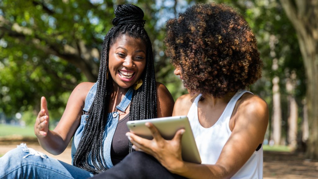 wo Black women are sitting in a park; one is turned to her friend smiling as her friend shows her something on her iPad